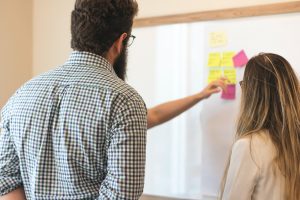 A man and a woman stand in front of a large whiteboard with sticky notes on it. Someone is explaining or teaching something on the board to the two people.